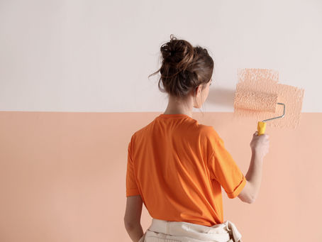 Woman in orange shirt painting a wall peach with a roller, standing in a brightly lit room.
