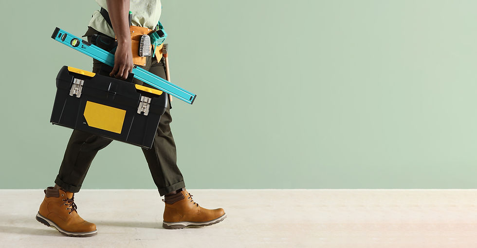 Person in work pants and boots walks holding a black toolbox with a yellow label and a turquoise level against a light green wall.