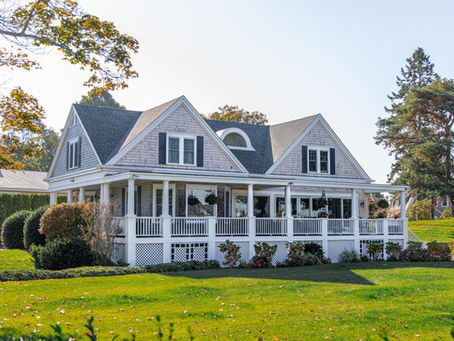 Two-story house with a gray roof, white porch, and manicured lawn surrounded by trees under a clear sky. Tranquil and inviting setting.
